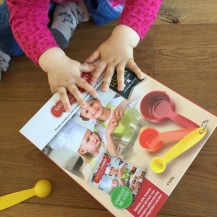 Auf dem Bild ist die Verpackung des Bechersets der kinderleichten Becherküche zu sehen. Ein Kleinkind hält die bunten Becher des Sets in der Hand.