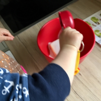 Ein Kind schüttet mit einem roten Becher der kinderleichten Becherküche Zucker in eine rote Schüssel.