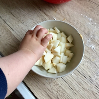 Das Bild zeigt wie eine kleine Hand Apfelstückchen aus einer Schüssel mit klein geschnittenen Apfelstückchen von einer bemehlten Arbeitsplatte nascht.
