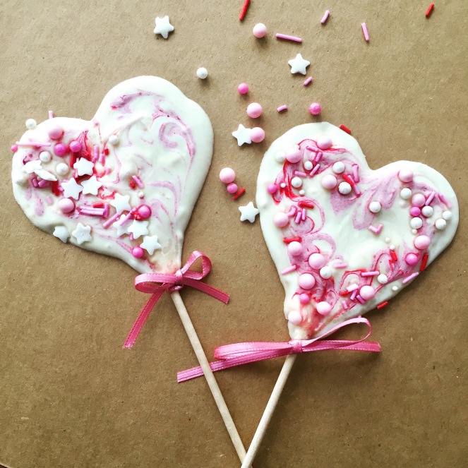 Auf Paperbraunem Hintergrund liegen zwei aus weißer Schokolade gegossene Herzlollis. Sie sind mit rosa eingefärbter Schokolade marmoriert und mit Zuckerperlen geschmückt.